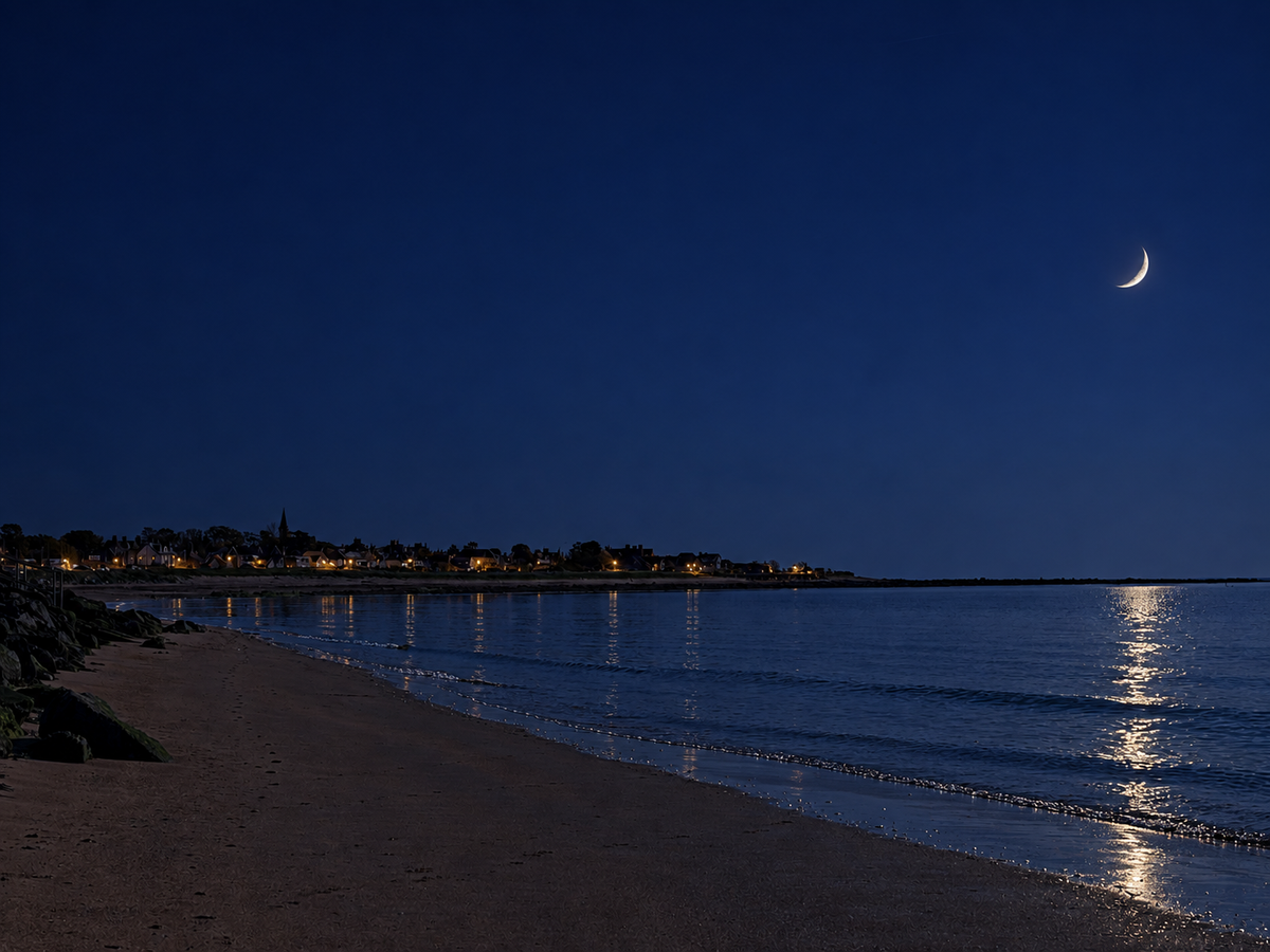 A serene night coastal scene with a sandy beach, calm water reflecting the moonlight, a distant shoreline with small warm lights, and a crescent moon in a deep blue sky. Original day time photo edited to night time by AI.